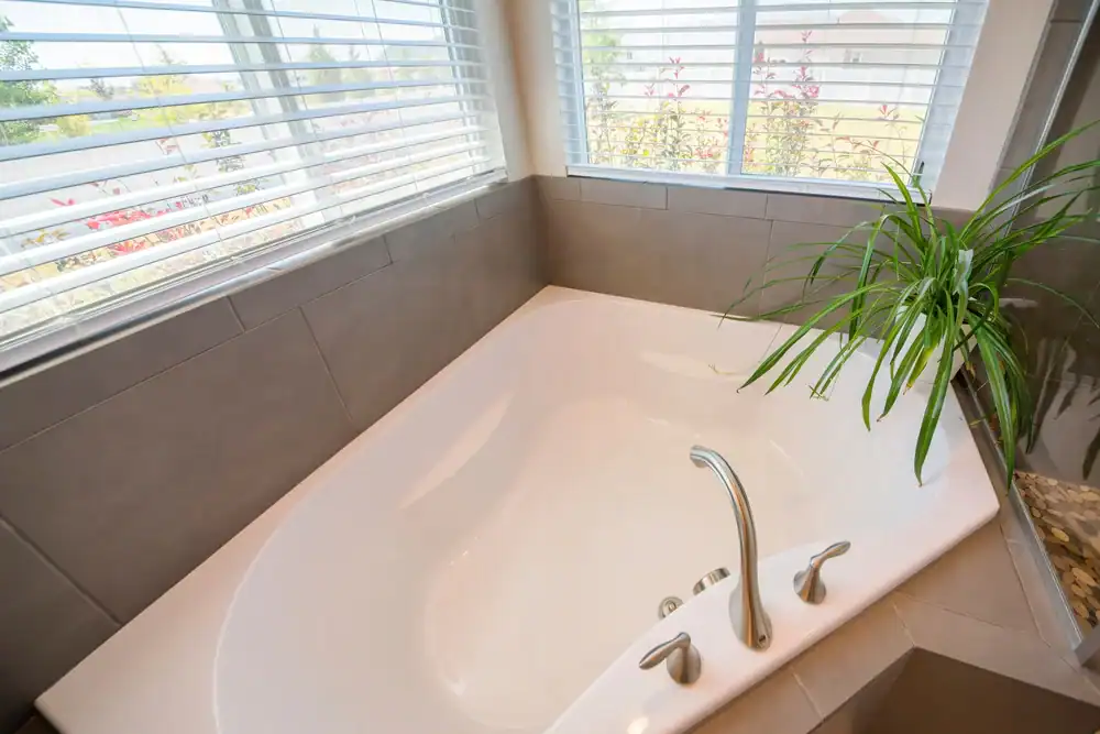 A modern white bathtub with silver faucets is set in a tiled corner beneath large windows with blinds. A green potted plant sits on the ledge, while sunlight pours in—perfect inspiration for marble restoration Nassau & Suffolk County, NY.