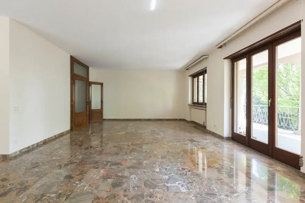 Spacious, empty room with marble flooring—recently enhanced by marble restoration in Nassau & Suffolk County, NY—large wooden-framed glass doors on one side, a window filling the white walls with natural light, and peaceful tree views outside.
