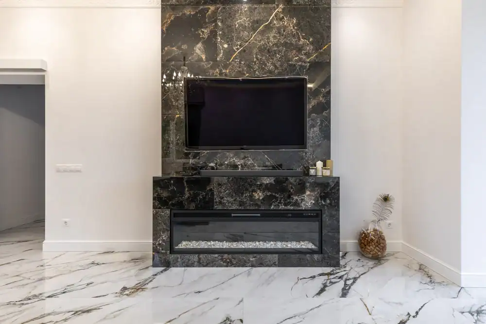 A modern living room features a black marble TV wall—with expert marble restoration Nassau & Suffolk County, NY—a mounted flat-screen TV, an electric fireplace below, white marble floors, and a decorative gold pineapple on the right.