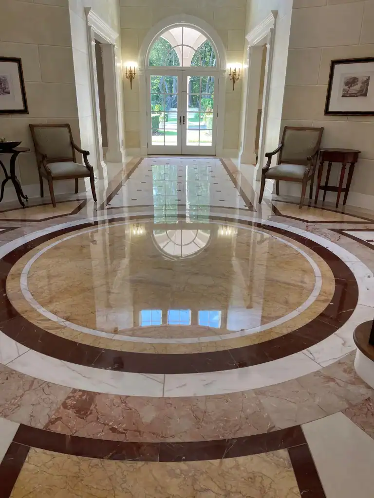 A polished marble floor, reflecting a large arched window and bright natural light, showcases expert marble restoration in Nassau & Suffolk County, NY. Two chairs and framed artwork accent the cream-colored walls of this elegant hallway.