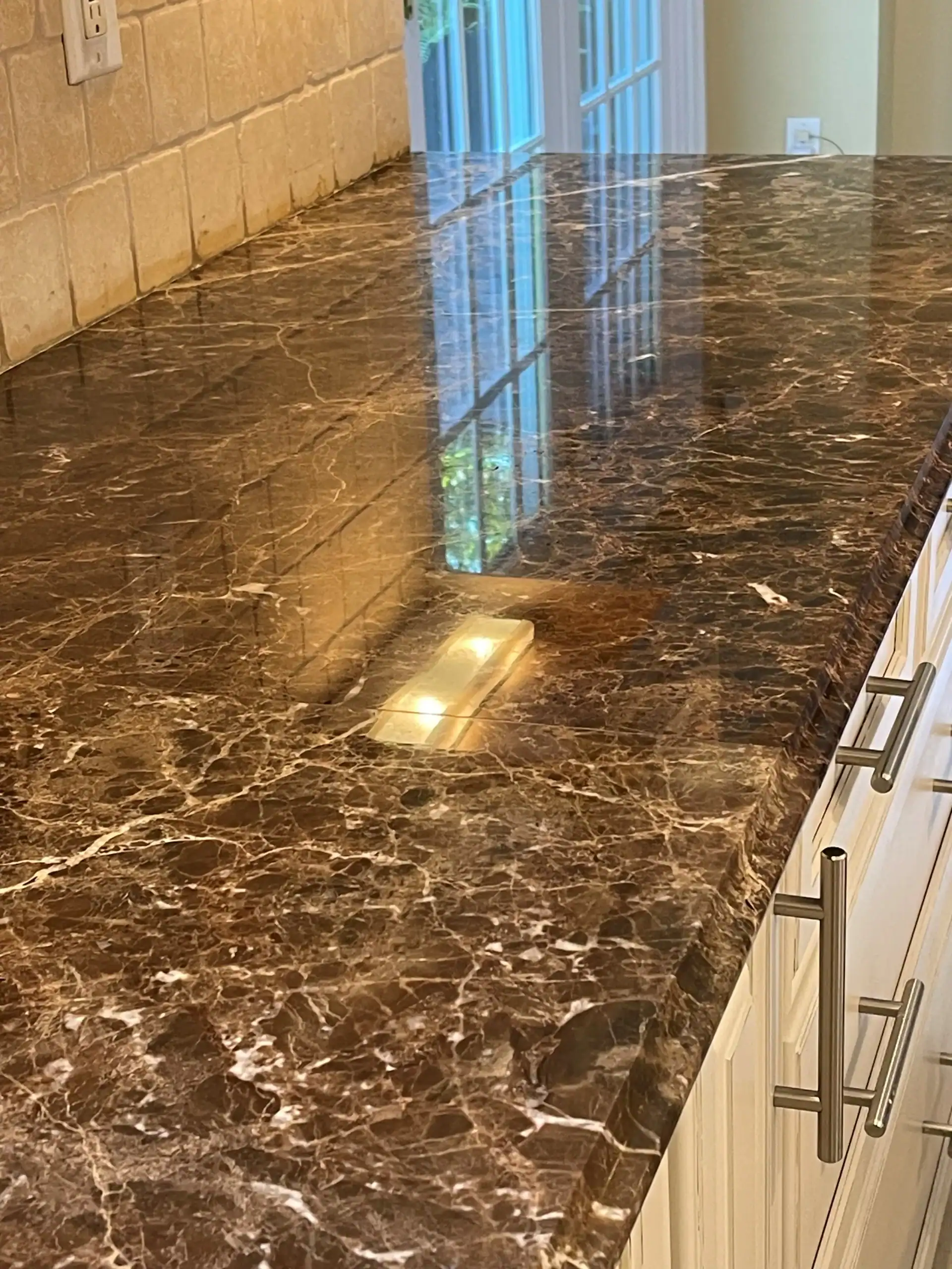 A close-up of a polished dark brown marble kitchen countertop with white veins, reflecting light and a window. Expert marble restoration in Nassau & Suffolk County, NY keeps the surface gleaming above white cabinets with silver handles.