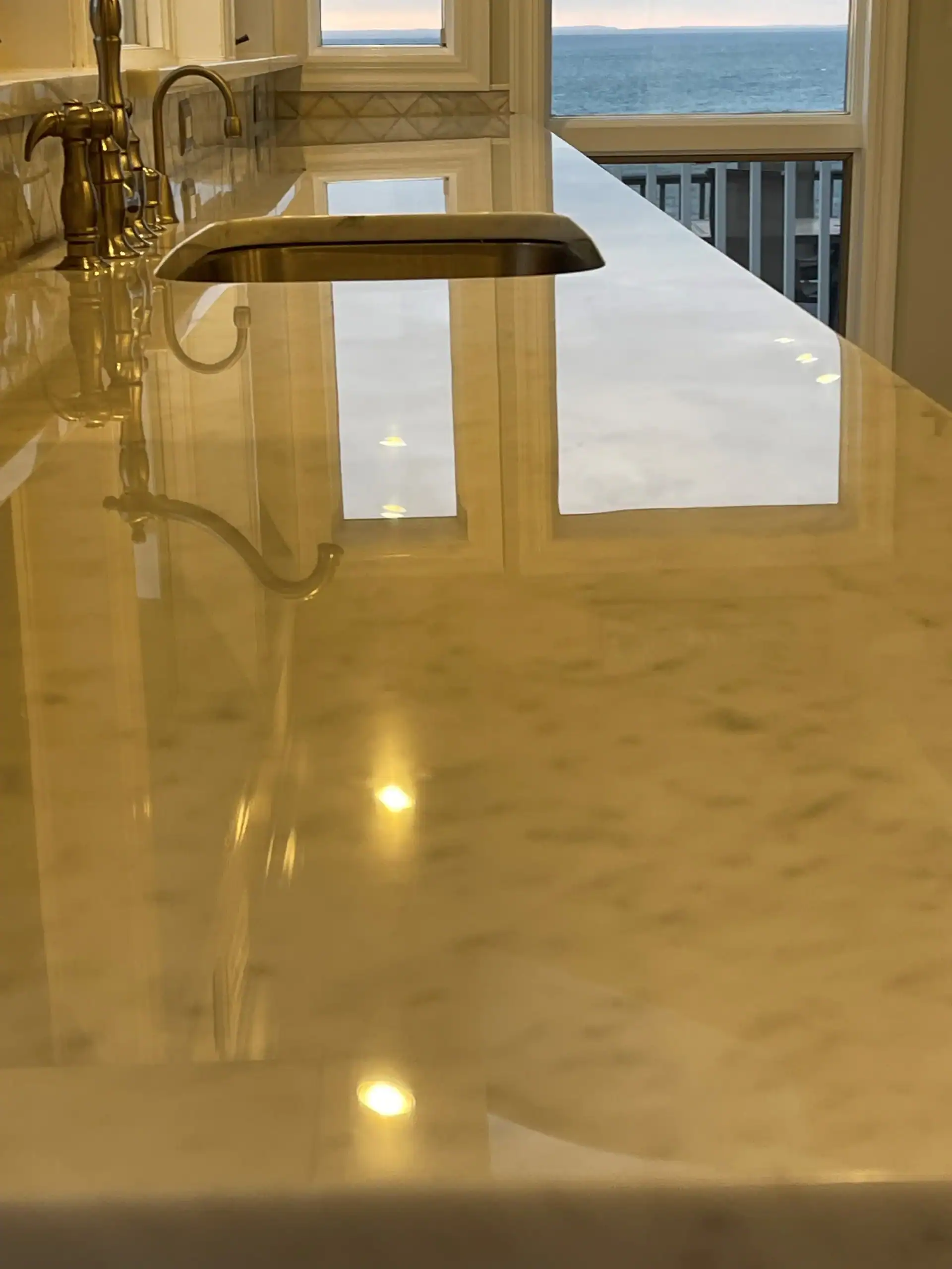 A shiny marble kitchen countertop with a built-in sink and faucet, reflecting overhead lights and a window view of a NY balcony and the ocean—perfectly maintained with expert marble restoration Nassau & Suffolk County services.