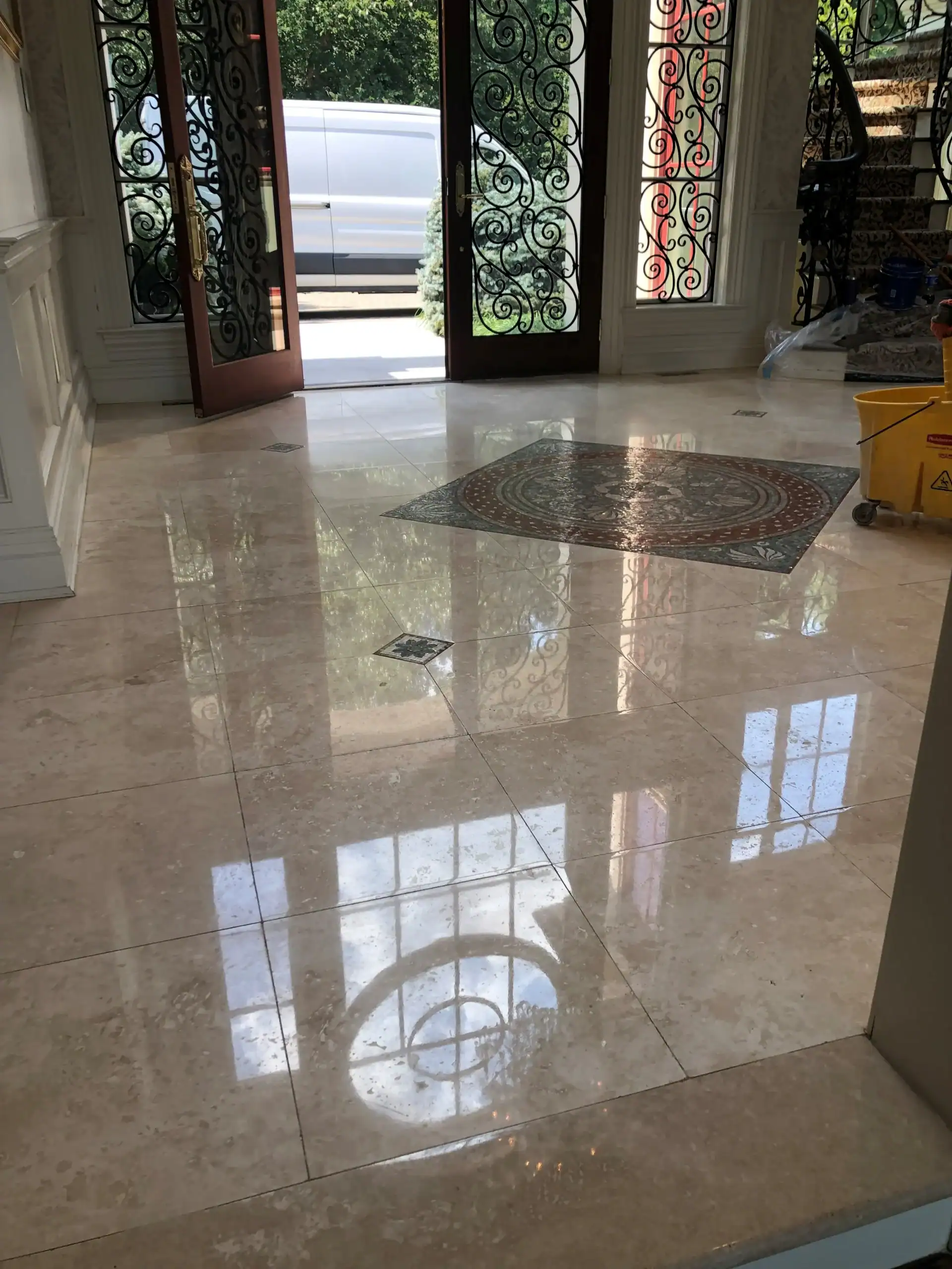 A shiny marble floor, expertly finished with marble restoration Nassau & Suffolk County, NY, reflects windows and decorative iron doors. Sunlight streams in from open doors leading to a leafy outdoor area and a parked car.
