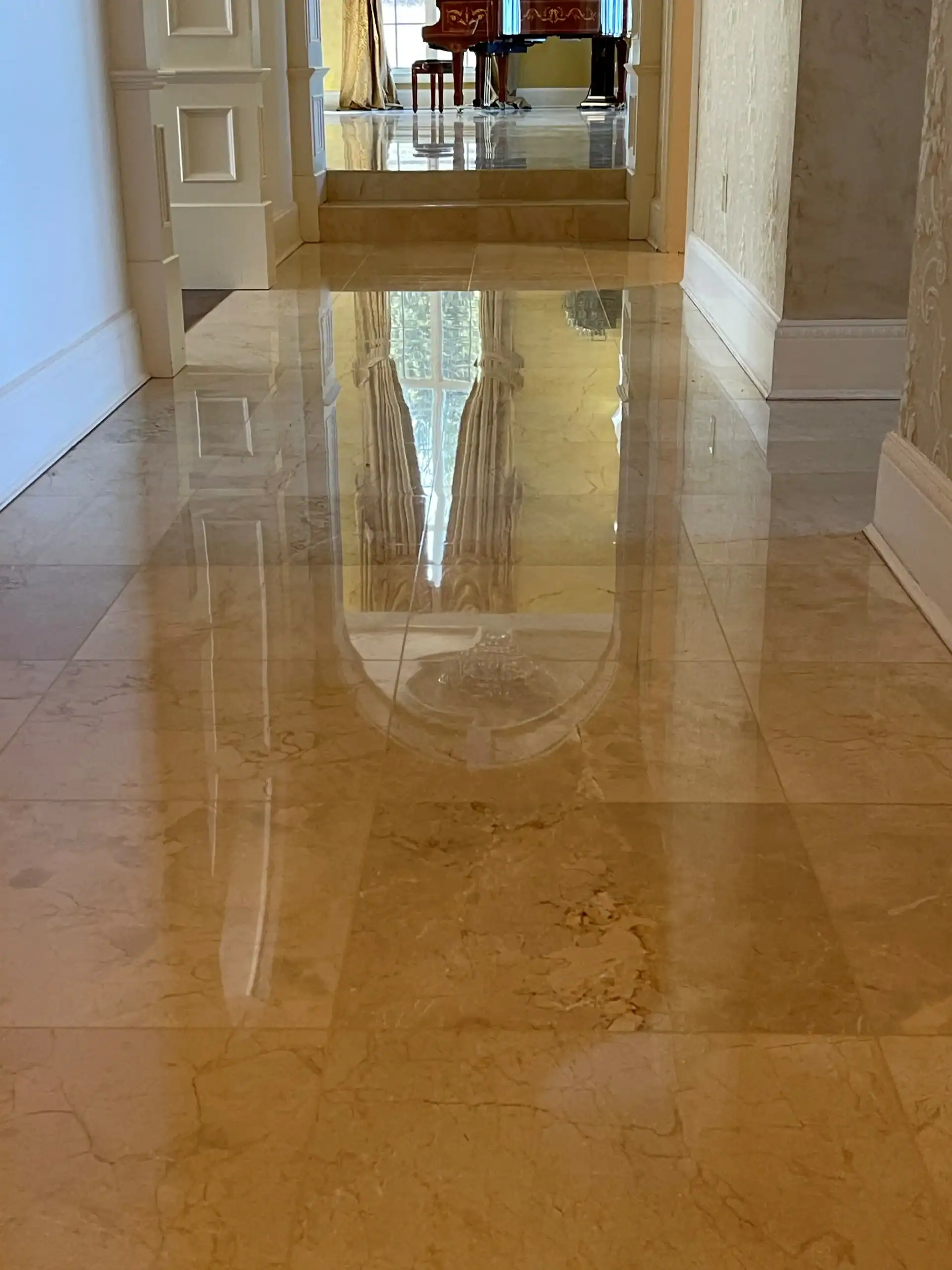 A highly polished marble hallway floor, enhanced by expert marble restoration in Nassau & Suffolk County, NY, reflects the light and nearby curtains, leading to a doorway with furniture and a piano visible in the background.