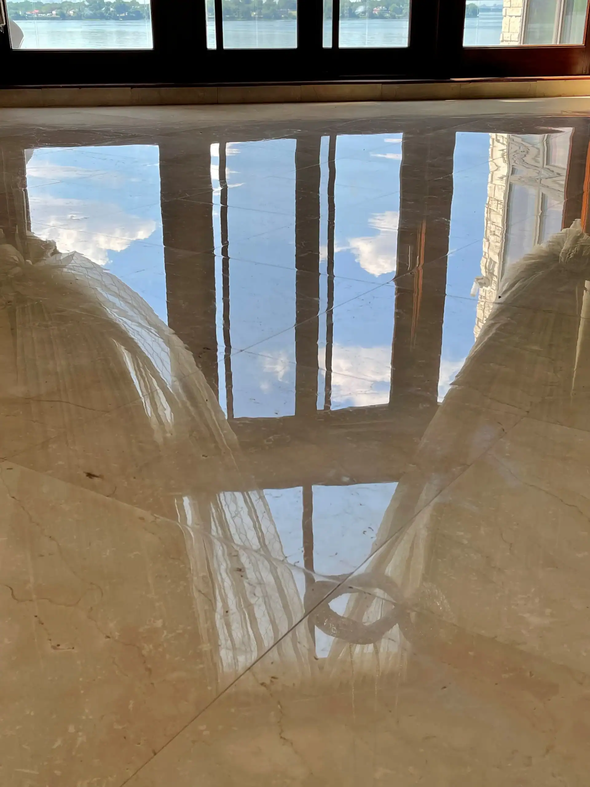 Sunlight streams through tall windows, reflecting blue sky and clouds on a shiny, newly finished floor—thanks to expert marble restoration in Nassau & Suffolk County, NY. Two sheer curtains frame the watery illusion on either side.