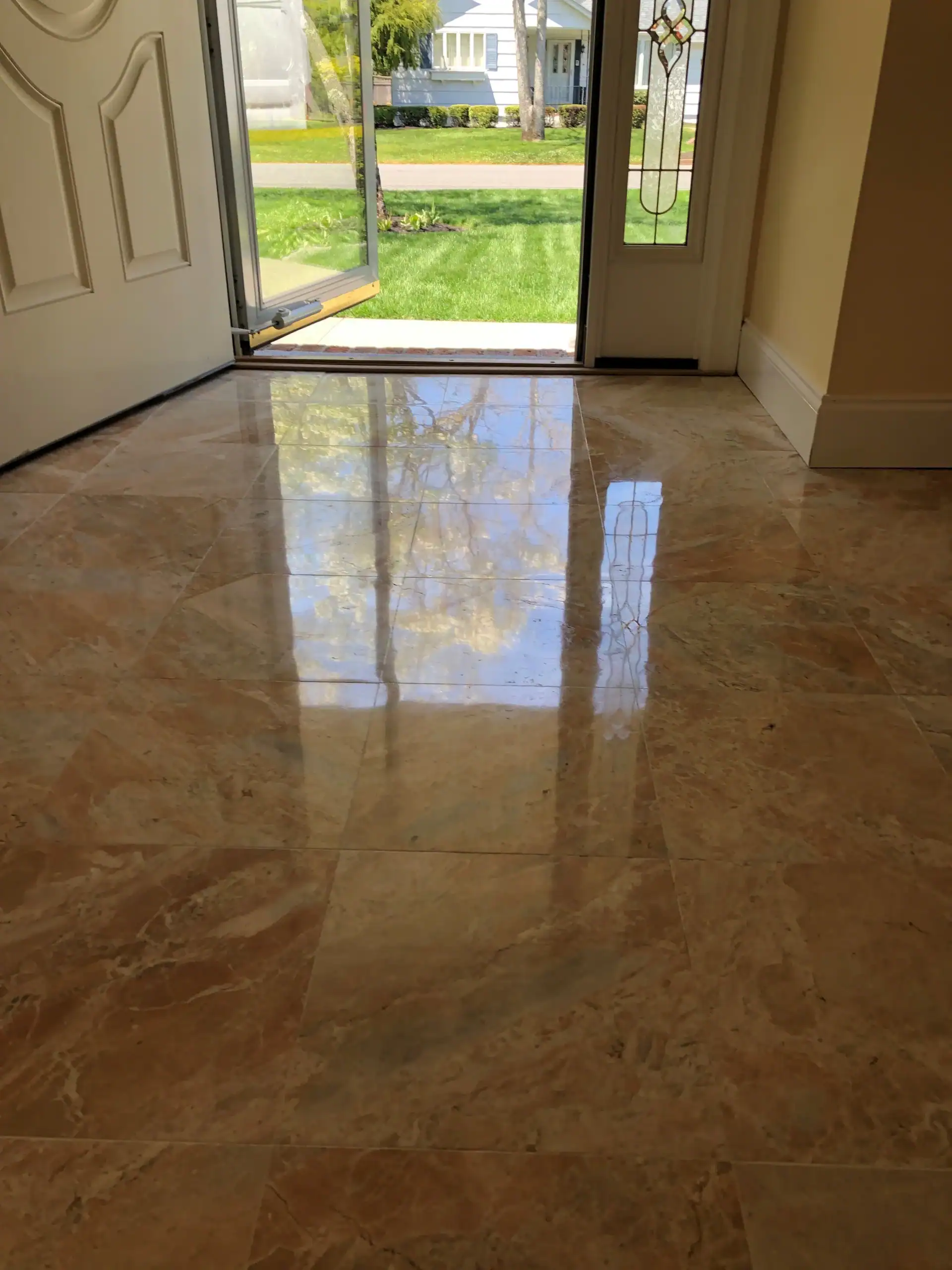 Sunlight and outdoor greenery reflect on shiny, polished marble floor tiles inside an open doorway, showcasing expert marble restoration Nassau & Suffolk County for a bright and welcoming NY home entrance.