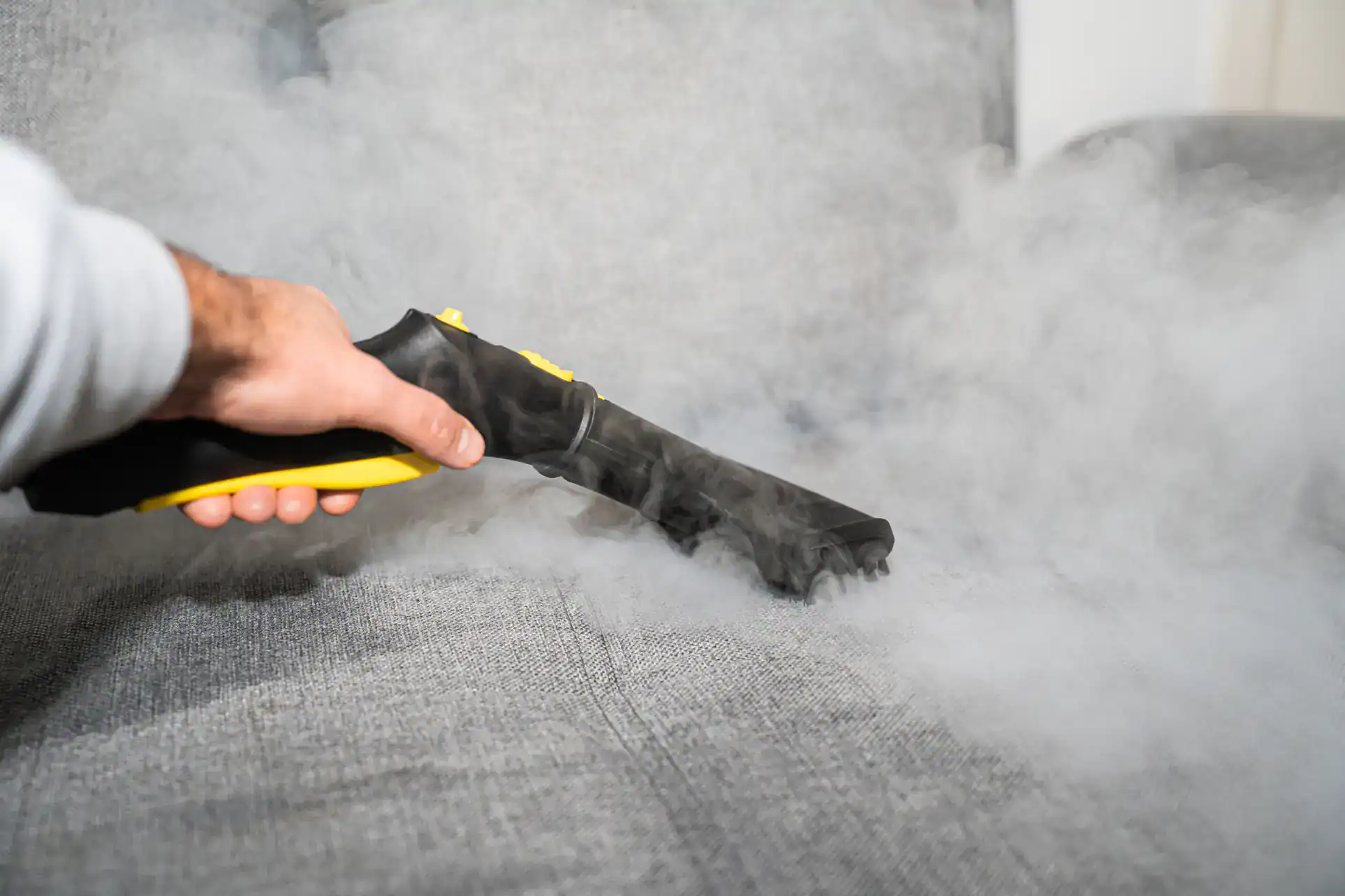 A person uses a handheld steam cleaner on a gray upholstered sofa, releasing visible steam onto the fabric for cleaning or sanitizing purposes.