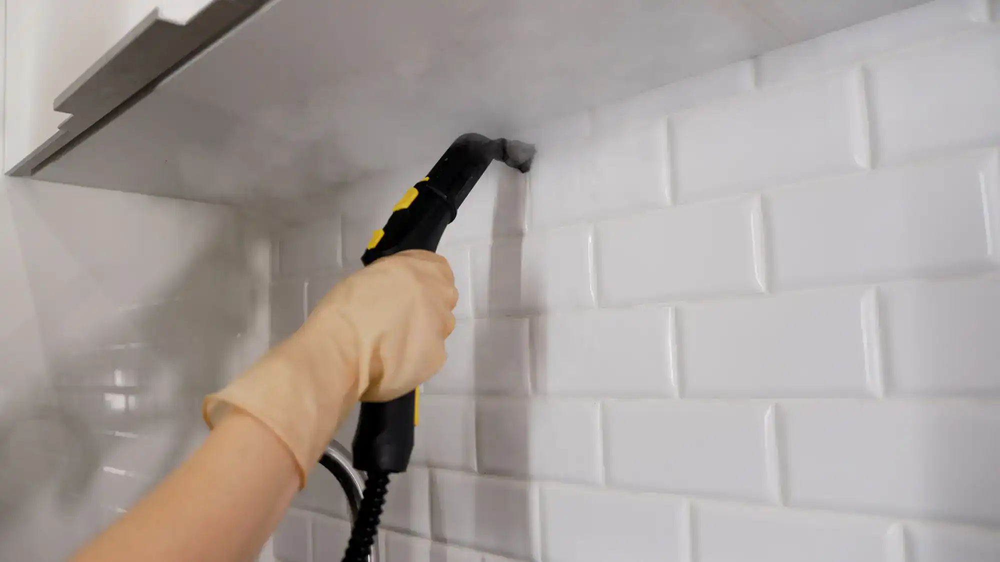 A person wearing a rubber glove uses a handheld steam cleaner to clean white subway tile backsplash under a kitchen hood, with visible steam coming from the nozzle.