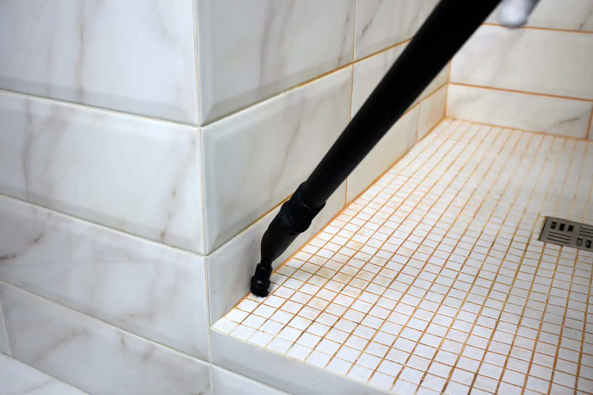 A steam cleaner nozzle is being used to clean the grout lines between tiles in a shower, removing dirt and stains from the white tiled surface and walls.