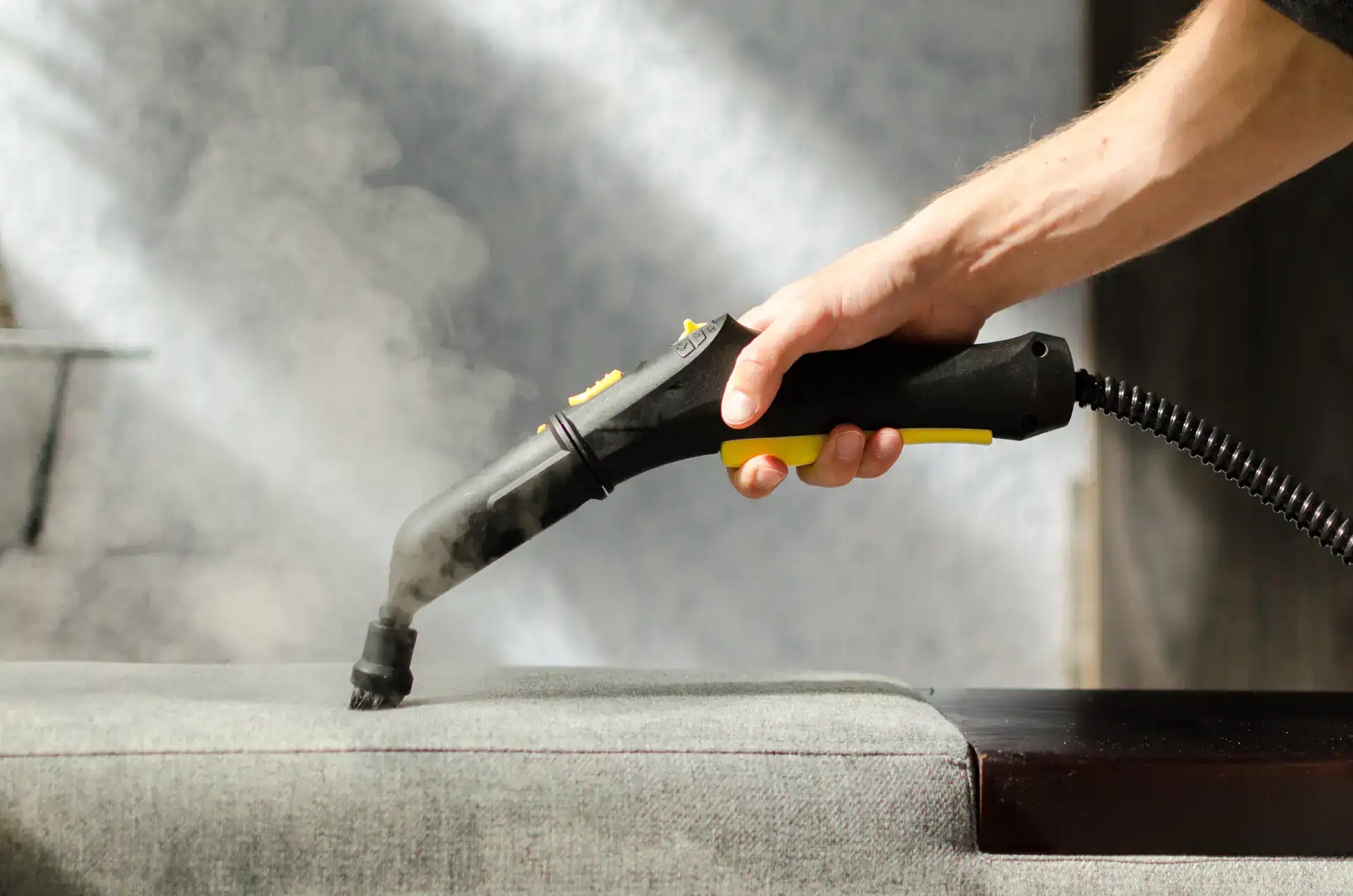 A hand holds a steam cleaner, releasing steam onto a gray upholstered surface, likely for cleaning or sanitizing the fabric.