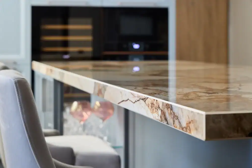 A close-up of a polished marble kitchen island countertop with a natural brown and cream pattern, accompanied by gray upholstered chairs and built-in appliances in the blurred background.