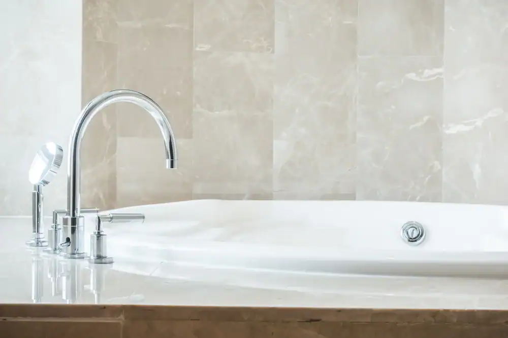 A modern bathtub with a shiny chrome faucet and handle, set against a light beige marble tiled wall and surrounded by a matching marble surface.