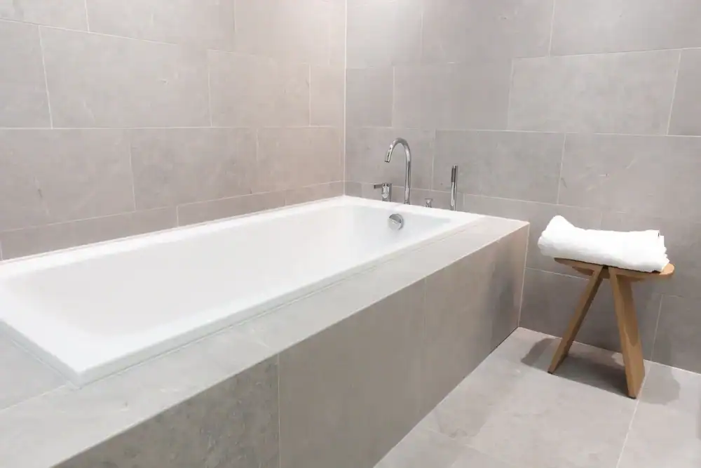 A modern bathroom with a built-in white bathtub surrounded by light grey tiles. Next to the tub is a small wooden stool holding a neatly folded stack of white towels.