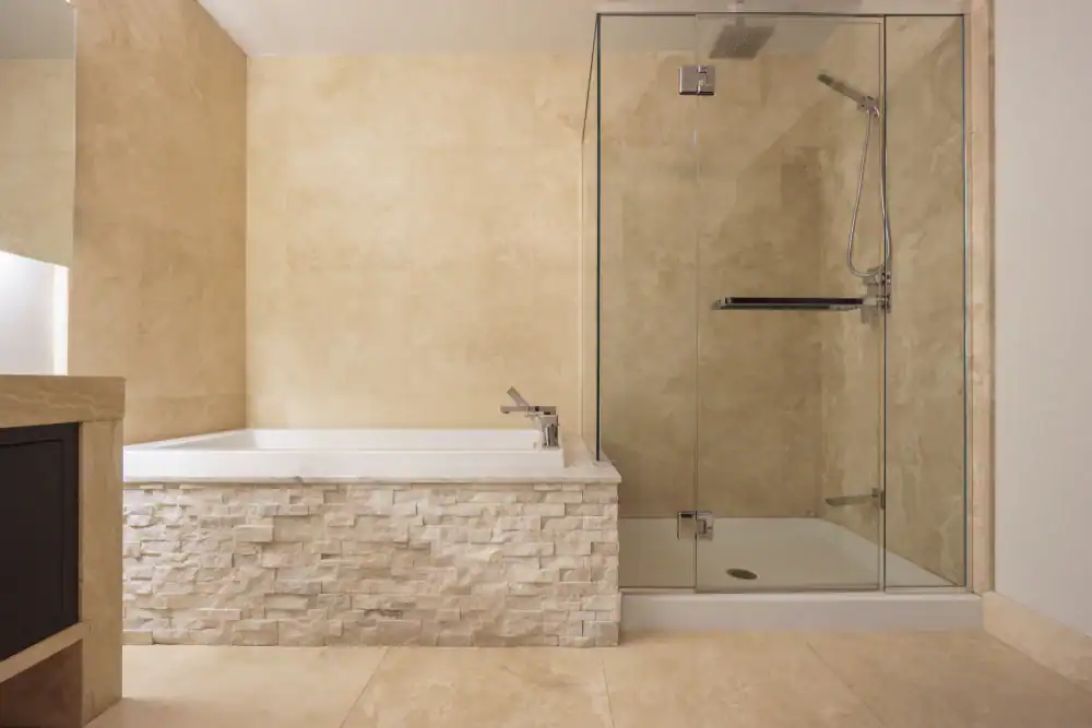 Modern bathroom with beige stone tiles, featuring a built-in bathtub with a stone fa&ccedil;ade on the left and a glass-enclosed shower with chrome fixtures on the right. The space looks clean and minimalistic.