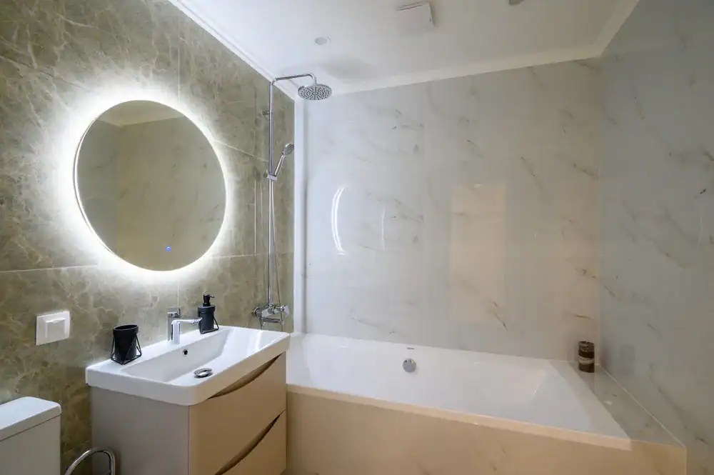 Modern bathroom with a round backlit mirror above a white sink, a bathtub with a wall-mounted shower, marble-style wall tiles, and minimalistic decor.