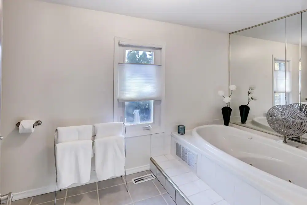 Modern bathroom with a large white bathtub, two white towels on a rack, a window with a shade, a toilet paper holder, and a large mirror above the tub. Decorative vases with flowers sit on the tub&rsquo;s ledge.