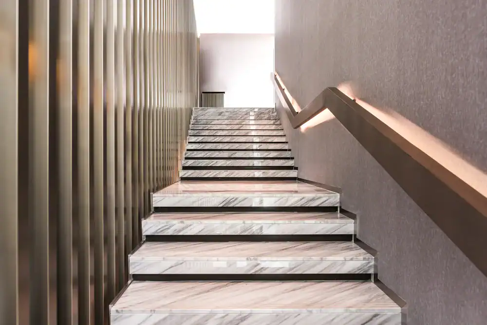 Marble staircase with a modern design, featuring a textured gray wall, sleek metal railing with built-in lighting on one side, and vertical gold-colored slats on the other side.