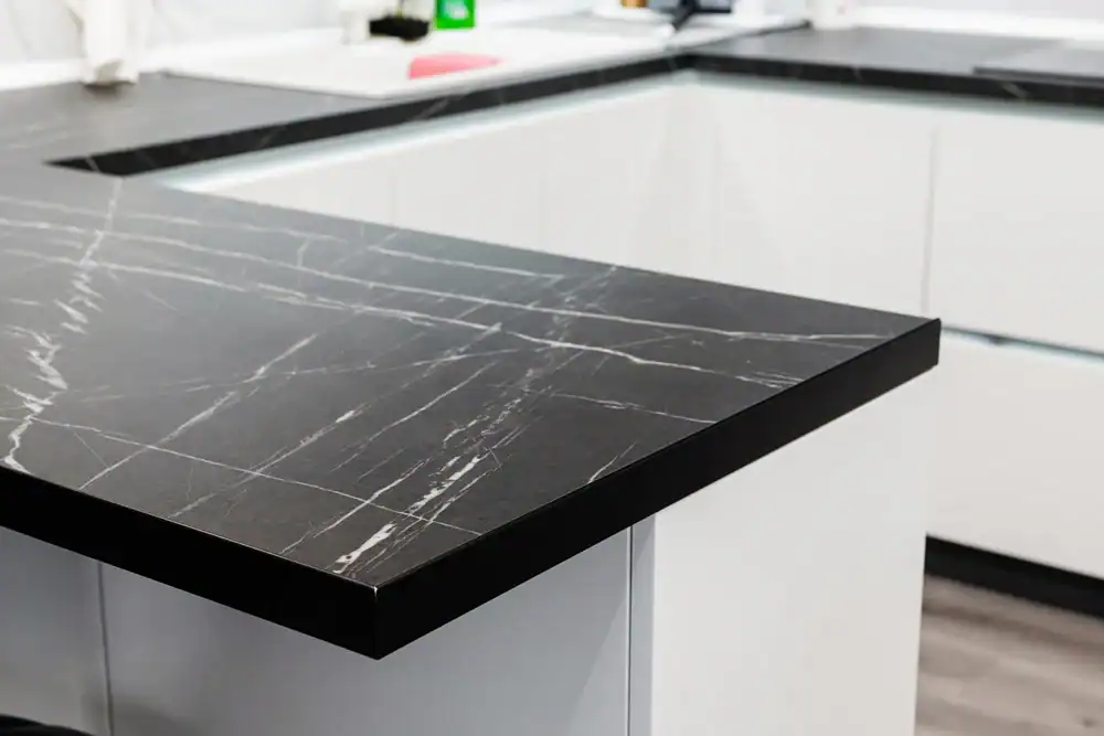 Close-up of a modern kitchen with a black marble countertop featuring white veining, set on white cabinets with minimalist handles. The countertop has a polished finish and the kitchen appears clean and contemporary.