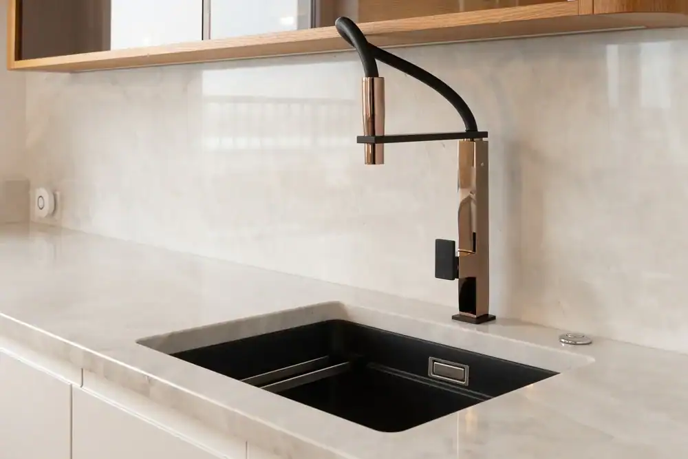 A modern kitchen countertop with a sleek black and gold faucet over a rectangular black sink, set against a light-colored marble backsplash and cabinets with wooden trim above.