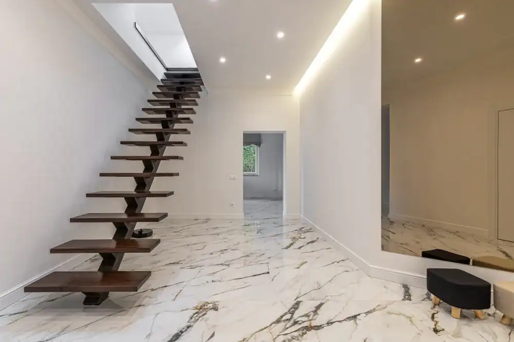 A modern interior with floating dark wooden stairs, white walls, recessed ceiling lights, and glossy marble flooring. A large wall mirror and two small stools are on the right side of the spacious room.