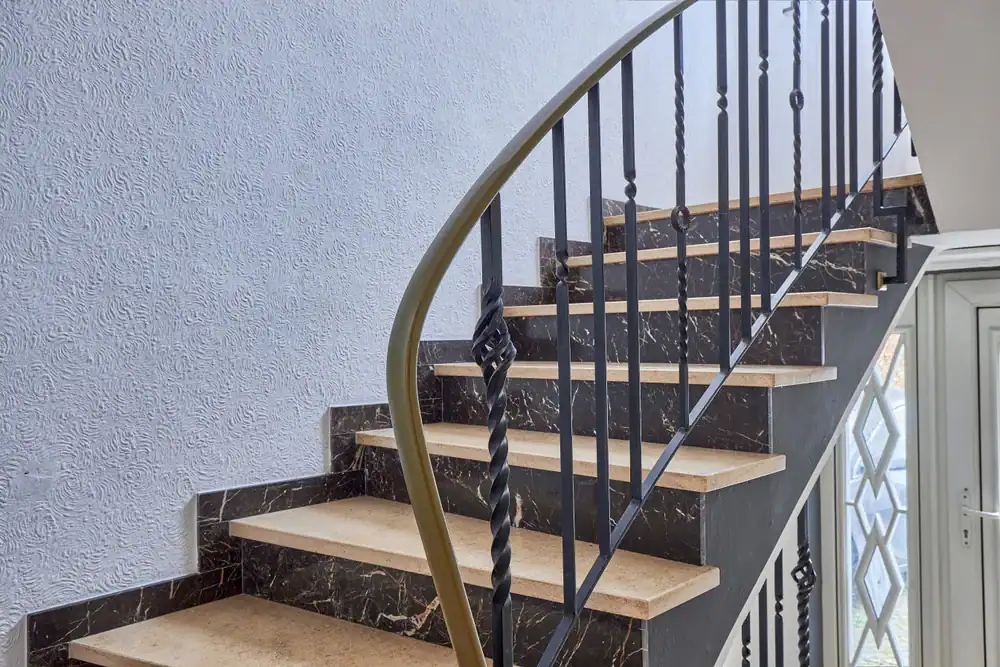 A modern staircase with beige marble steps, black marble risers, and a black metal railing with decorative twists, positioned beside a textured white wall. Natural light enters from nearby windows.